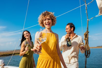 Drei fröhliche Menschen auf einem Boot, die bei Sonnenschein mit Sektgläsern anstoßen und lachen.