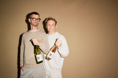 Zwei Männer stehen mit jeweils einer Flasche haltend vor ausgeleuchteten ockerfarbenen Wand