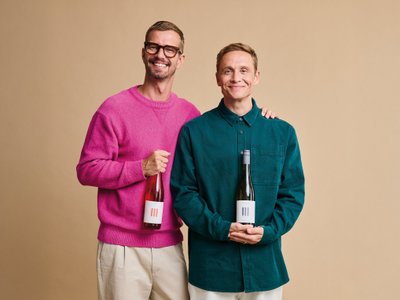 Zwei Männer stehen lächelnd nebeneinander und halten Flaschen mit Wein in der Hand, vor einem neutralen Hintergrund.